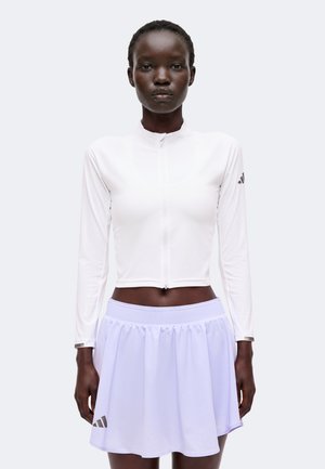 Γυναίκα που φοράει λευκό μακρυμάνικο crop top με φερμουάρ και λευκή πλισέ αθλητική φούστα σε απλό φόντο.