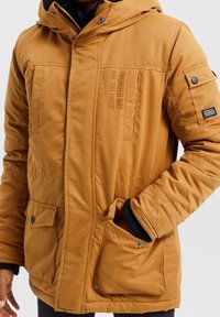 Honigfarbene, isolierte Jacke mit Kapuze, ausgestattet mit Fronttaschen, verstellbaren Manschetten und einem bedruckten Logodetail auf der Brust.