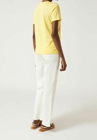 Žluté bavlněné tričko s krátkým rukávem, spárované s bílými kalhotami rovného střihu a hnědými plochými sandály. Jednoduchý design, bez viditelných vzorů.