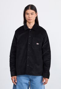 Camisa de pana negra con botones en el frente, cuello, bolsillo en el lado izquierdo del pecho con un parche de logo. Mangas largas con botones en los puños.