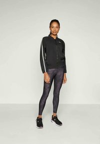 Sort atletisk jakke med hvide striber, parret med sort mønstrede leggings. Modellen har sorte sneakers på med pink detaljer.