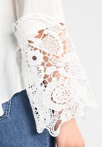 Haut blanc avec des manches en dentelle complexe aux motifs floraux. La dentelle présente une texture délicate et un design ajouré, associée à un corps en tissu lisse.