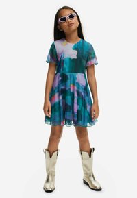 Kleurvolle mouwloze jurk met een bloemenontwerp in teal, paars en roze. Heeft een uitlopende silhoeët en geschulpte mouwen. Gecombineerd met zilveren cowboylaarzen.