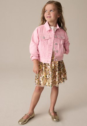 Fată tânără cu părul lung, purtând o jachetă roz din denim, o fustă cu paiete aurii și pantofi Mary Jane aurii, stând cu o mână în buzunar.