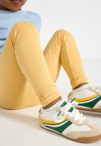 Barn sidder med bøjede knæ, iført gule leggings, hvide sneakers med grønne og gule striber og hvide sokker på et lyst gulv.
