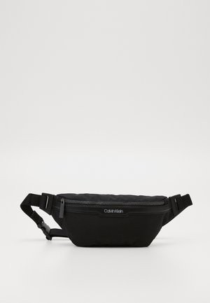 Sort Calvin Klein bæltetaske med justerbar rem og lynlåslukning, vist mod en ensfarvet hvid baggrund.
