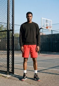 Jeune homme en sweat-shirt noir et short rouge debout sur un terrain de basket extérieur près de la clôture et du panier sous un ciel clair.