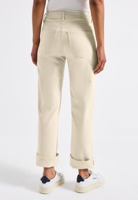 Pantalones rectos de mezclilla en color beige claro, con puños enrollados y dos bolsillos traseros. Llevados con zapatillas blancas.