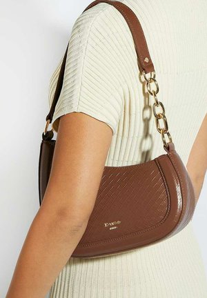 Sac bandoulière en cuir marron tressé avec chaînes en or porté au bras par-dessus un haut crème côtelé à manches courtes.