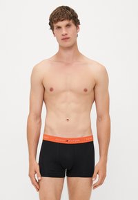 Schwarze Boxershorts mit einem roten Bund, der das "TOMMY HILFIGER"-Logo in Weiß zeigt. Das Material scheint glatt und körpernah zu sein.