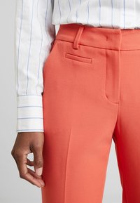 Oranžové kalhoty vyrobené ze strukturované látky s plochým předním dílem, bočními kapsami a poutky na opasek. V kombinaci s bílou košile s modrými pruhy.