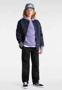 Marine Bomberjacke, lilafarbener Kapuzenpullover, schwarze Hose und schwarz-weiße Sneakers; ausgestattet mit Seitentaschen und einer hellblauen Kappe mit Logo.