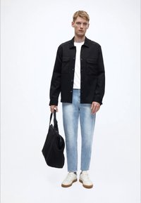 Svart skjorte med knapper foran og brystlommer, hvit t-skjorte, lyseblå jeans, hvite joggesko med beige såler og en svart skulderveske.