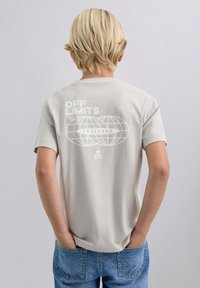 T-shirt en coton gris clair avec un grand graphique blanc représentant "INTERDIT" et un design de globe au dos. Manches courtes, coupe décontractée.