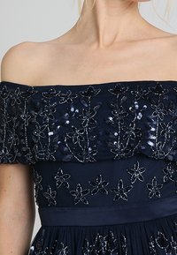 Šaty bez ramínek v námořnické modré barvě, zdobené květinovou vyšívkou z flitrů, s upnutou přední částí a plisovanou sukní. Měkká tkanina s hladkým povrchem.