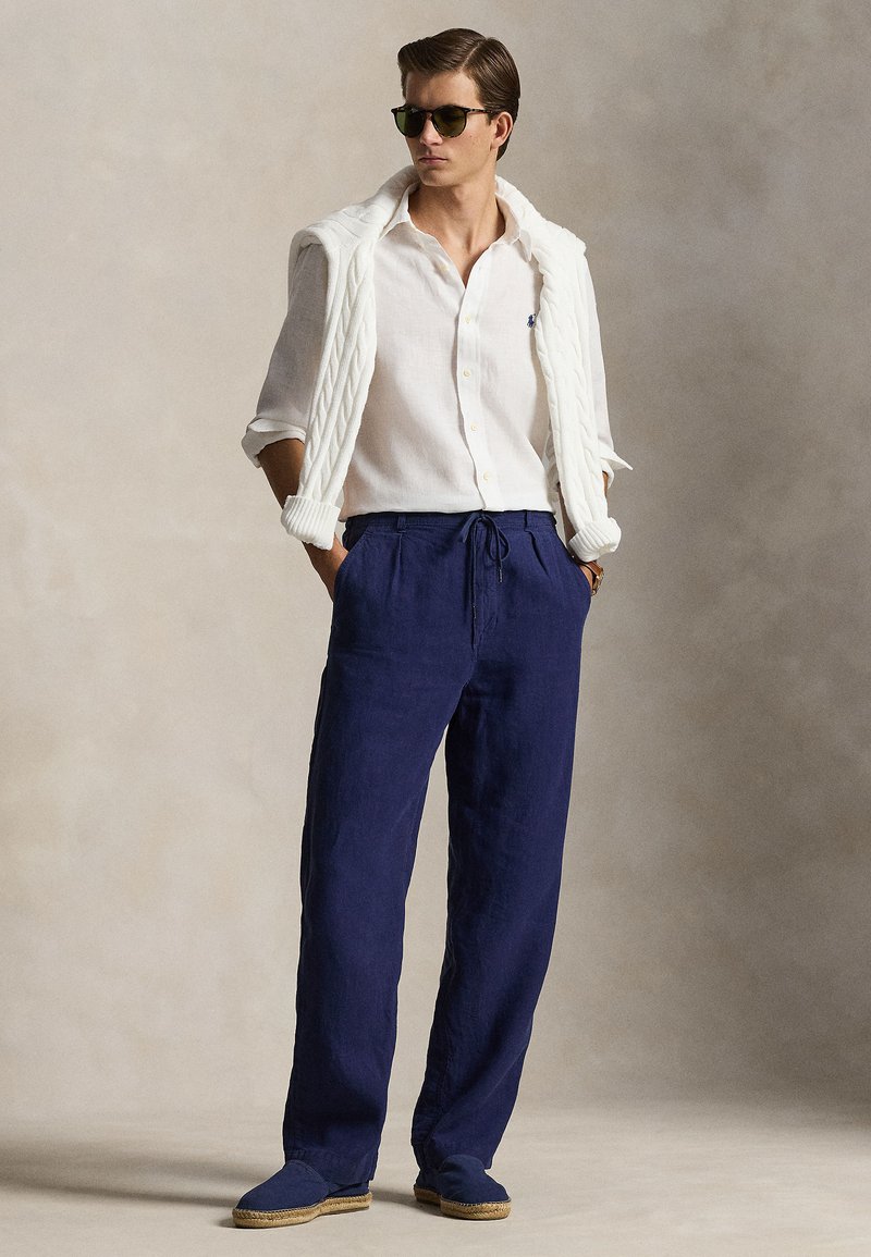 Mann in weißem Hemd mit Knöpfen und weißem Pullover, der über die Schultern gelegt ist, marineblaue Kordelzug-Hose, dunkle Sonnenbrille und Slipper.