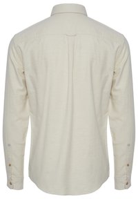 Chemise longue beige clair à manches longues avec un col standard, des poignets boutonnés, un détail de empiècement au dos et des accents de couture bleus subtils sur les manches.