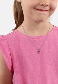 Collana in argento con pendente a forma di cuore che presenta un piccolo gemma, indossata sopra un top di tessuto rosa con maniche corte.