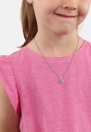 Collar de cadena de plata con un colgante en forma de corazón que presenta una pequeña gema, llevado sobre una blusa de tela rosa de mangas cortas.