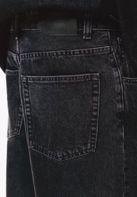 Gros plan sur un jean en denim noir montrant la poche arrière, la boucle de ceinture et les détails cousus sur la texture du tissu.