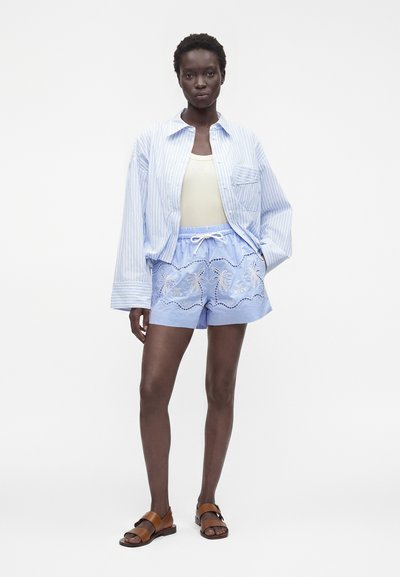 Femme portant une chemise oversize rayée légère, un débardeur blanc, un short brodé bleu clair et des sandales marron, debout devant un fond uni.