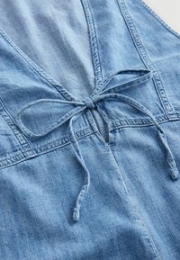 Vêtement en denim bleu clair avec un détail de lacet noué à l'avant et des coutures visibles sur tissu doux.