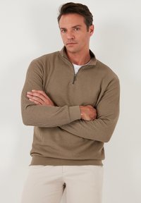 Pull beige zippé avec texture côtelée, manches longues et col montant, porté sur une chemise blanche, associé à un pantalon de couleur claire.