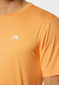 Oranžs sporta krekls, kas izgatavots no gludas, elastīgas auduma. Tam ir apaļa kakla izgriezums un mazs balts logotips kreisajā augšējā krūtīs.
