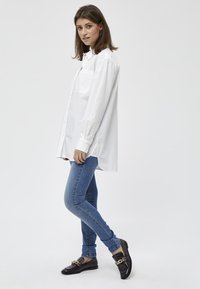 Camicia bianca con bottoni e maniche lunghe, tasca frontale, abbinata a jeans skinny blu e mocassini neri con un dettaglio in catena dorata.