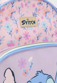 Ροζ υφασμάτινο σακίδιο με floral σχέδιο και μπλε φερμουάρ, με ετικέτα "STITCH" και διακοσμητικά με χαρακτήρα κινουμένων σχεδίων στο μπροστινό μέρος.