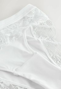 Tissu blanc orné de dentelle avec motifs floraux et détails en maille fine, montrant un gros plan de couches textiles douces et délicates.