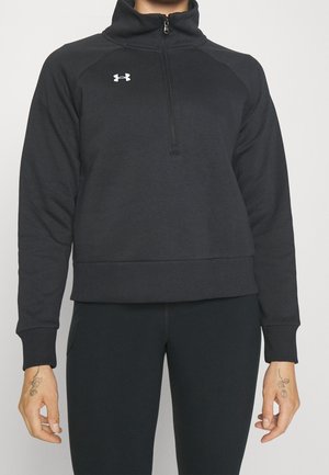 Personne portant un sweat-shirt noir Under Armour à fermeture éclair quart-de-tour et des leggings noirs, avec de petits tatouages floraux sur les deux mains.