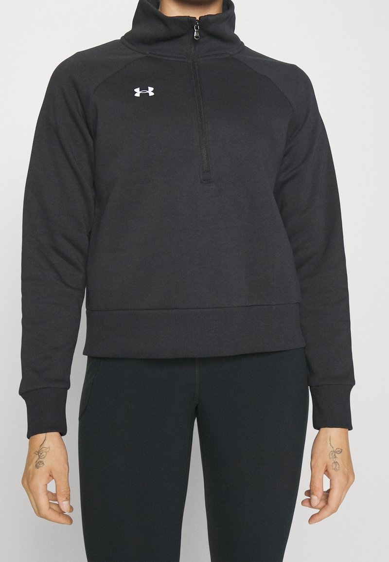 Personne portant un sweat-shirt noir Under Armour à fermeture éclair quart-de-tour et des leggings noirs, avec de petits tatouages floraux sur les deux mains.