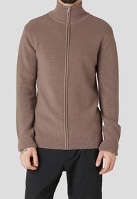 Brauner, gerippter Strickpullover mit durchgehendem Reißverschluss und hohem Kragen, der über einer dunklen Hose getragen wird.