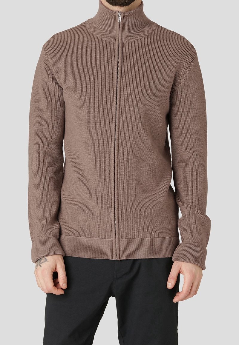 Brauner, gerippter Strickpullover mit durchgehendem Reißverschluss und hohem Kragen, der über einer dunklen Hose getragen wird.