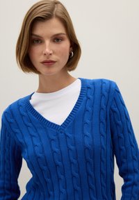 Blauwe gebreide trui met een V-hals en een gestructureerd kabelpatroon. Lange mouwen, gedragen over een wit shirt, wat contrasterende kleuren toont.