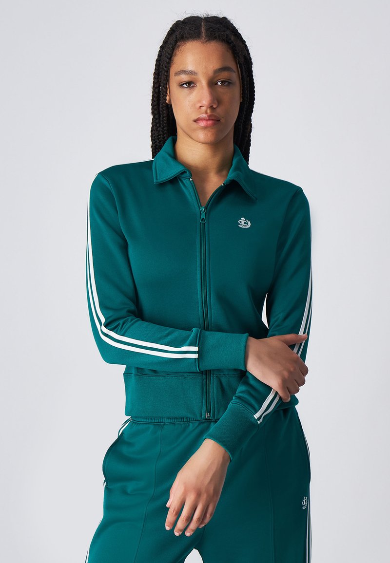 Chaqueta de chándal azul verdoso con detalles en blanco en las mangas, tela texturizada y un logo en el pecho. Diseño ajustado con cuello.