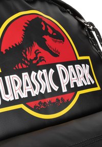 Bolsa negra con un logo rojo y amarillo de "Jurassic Park" que incluye la silueta de un dinosaurio y letras blancas, fabricada con material texturizado.