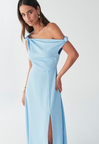 hele helesinine, õlgadest paljastav kleit sõlmitud varrukatega, põrandapikkusega, küljepealse lõhikuga ja siledast, voolavast kangast.