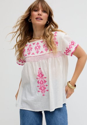 Frau trägt weiße locker sitzende Bluse mit rosa geometrischer Stickerei, blaue Jeans, goldene Creolen und goldenes Armband vor einfachem Hintergrund.