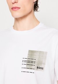 T-shirt en coton blanc avec un graphique carré texturé olive et des lignes noires, avec "BOSS" imprimé en gras en bas à droite.