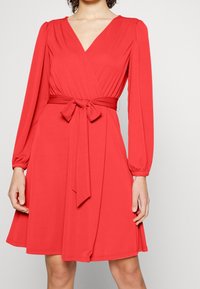 Červené šaty s dlouhými rukávy, výstřihem do V, úzkým pasem a odpovídajícím páskem ze stejného materiálu. Hladká textura a rozšířená sukně.