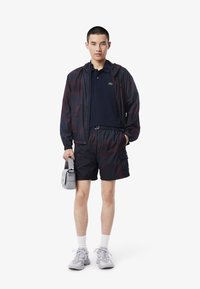 Voděodolná, tmavě červená a černá bunda, námořnické polo, černé cargo šortky, šedé tenisky a malá šedá taška, s lehkým designem.