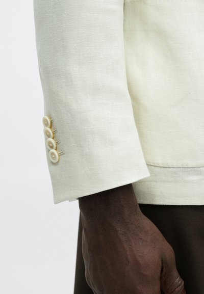 Main à la peau foncée à côté d'une manche de veste crème avec quatre boutons beige, portant un pantalon marron foncé sur fond blanc.
