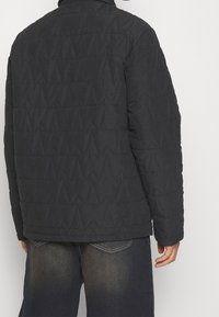 Black quilted jacket featuring a geometric pattern, round collar, and long sleeves, paired with dark denim jeans.