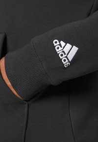 Czarna tkanina bluzy z prążkowanymi ściągaczami. Na lewym rękawie znajduje się biały haftowany logo Adidas. Gładka tekstura.