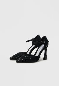 Μαύρα ψηλοτάκουνα παπούτσια από καστόρι με μυτερή μύτη, λουράκι στον αστράγαλο, διακοσμημένα με μικρά τρουκς στο μπροστινό μέρος και μοναδικό σχέδιο τακουνιού τύπου block.