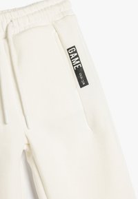 Hvide sweatpants lavet af blødt bomuldsblanding, med elastisk talje, snøre og et sort mærke med "GAME" på siden.