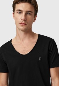 Camiseta de algodón negra con un diseño de escote en V profundo, que presenta un pequeño bordado de cráneo de cabra gris en el pecho izquierdo. Textura suave.