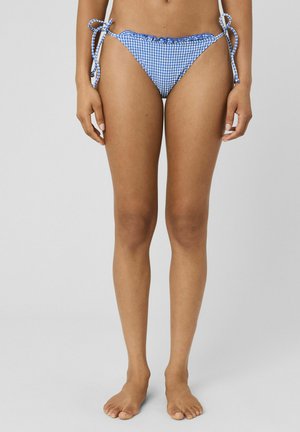 Femme portant un bas de bikini à carreaux bleu et blanc avec des liens sur les côtés, debout pieds nus sur un fond gris uni.